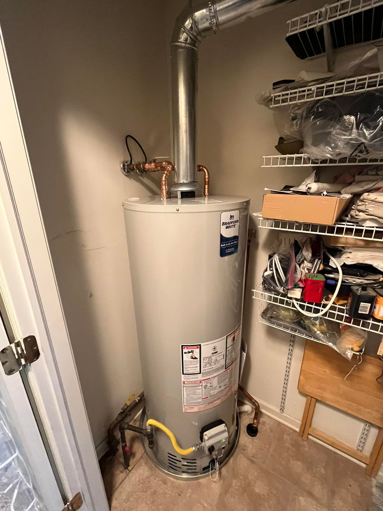 A water heater is sitting in a closet next to a shelf.