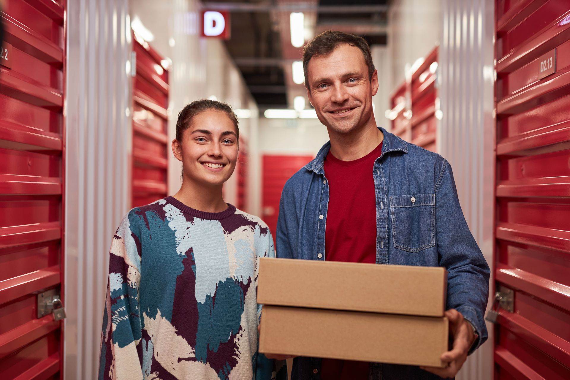 Two people standing in a storage facility hallway, holding cardboard boxes.