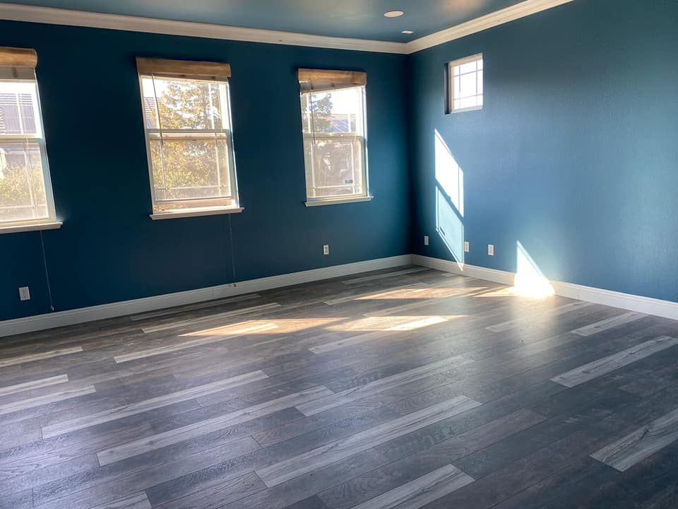 Empty room with teal walls, grey wood-look floor, windows with shades, white trim. Sunlight streaks across the floor.