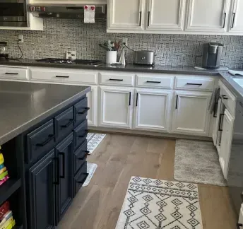 Kitchen with white cabinets, black hardware, dark island, and patterned rugs. 