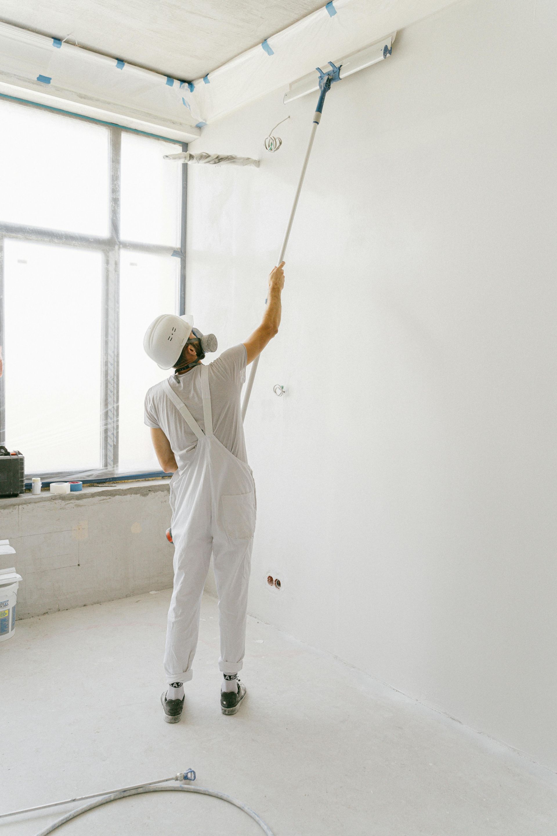 Person in white protective suit spraying paint on a white wall in a room.