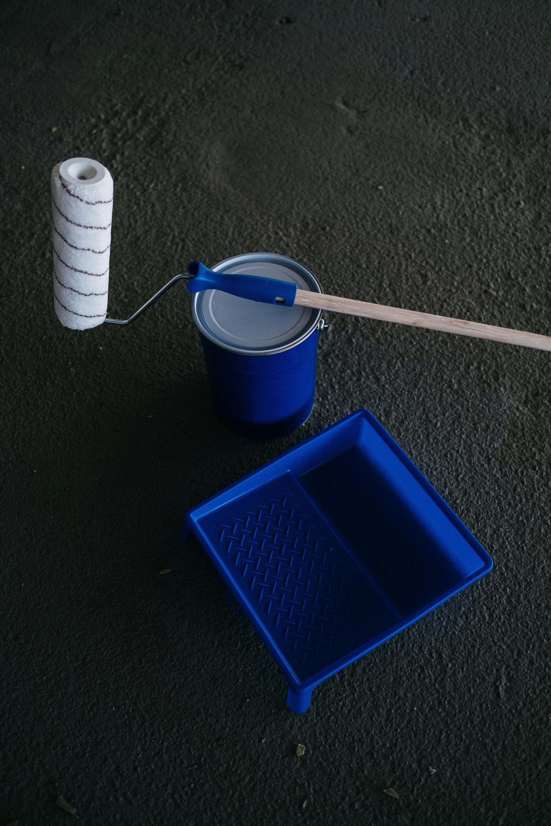 Paint roller, blue paint can, and tray on a dark surface, ready for painting.