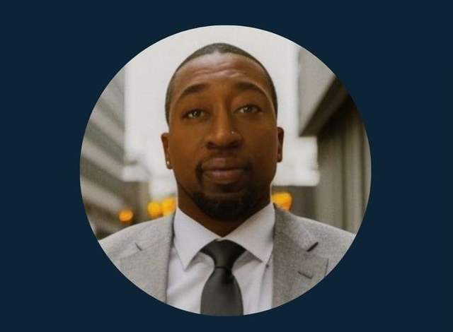 Woman sitting at a desk, holding a mug, looking at the viewer. A laptop, phone and window are visible.