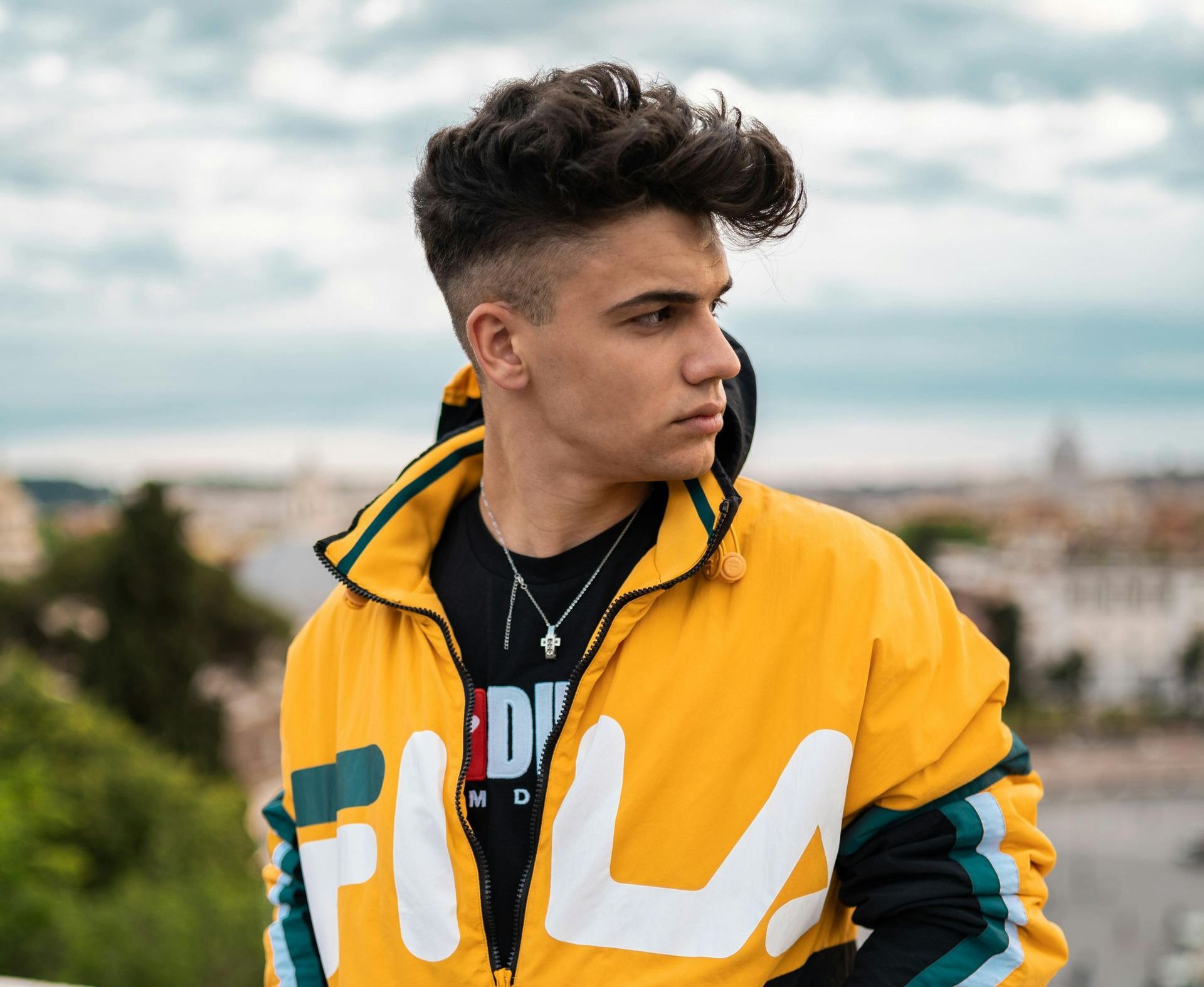 Young person wearing yellow FILA jacket looks away from the camera, standing outdoors with a cityscape background.