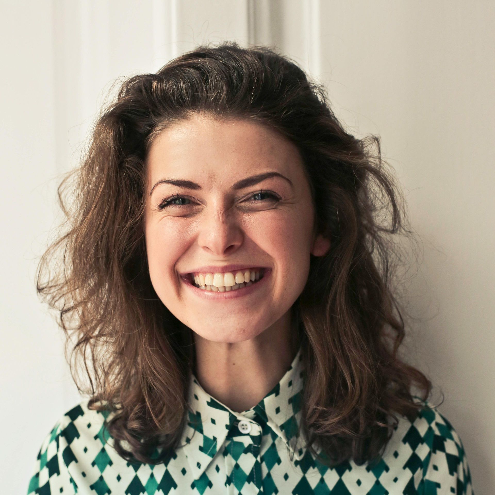 Woman with curly brown hair smiles broadly, wearing a green geometric patterned shirt in front of a white door.