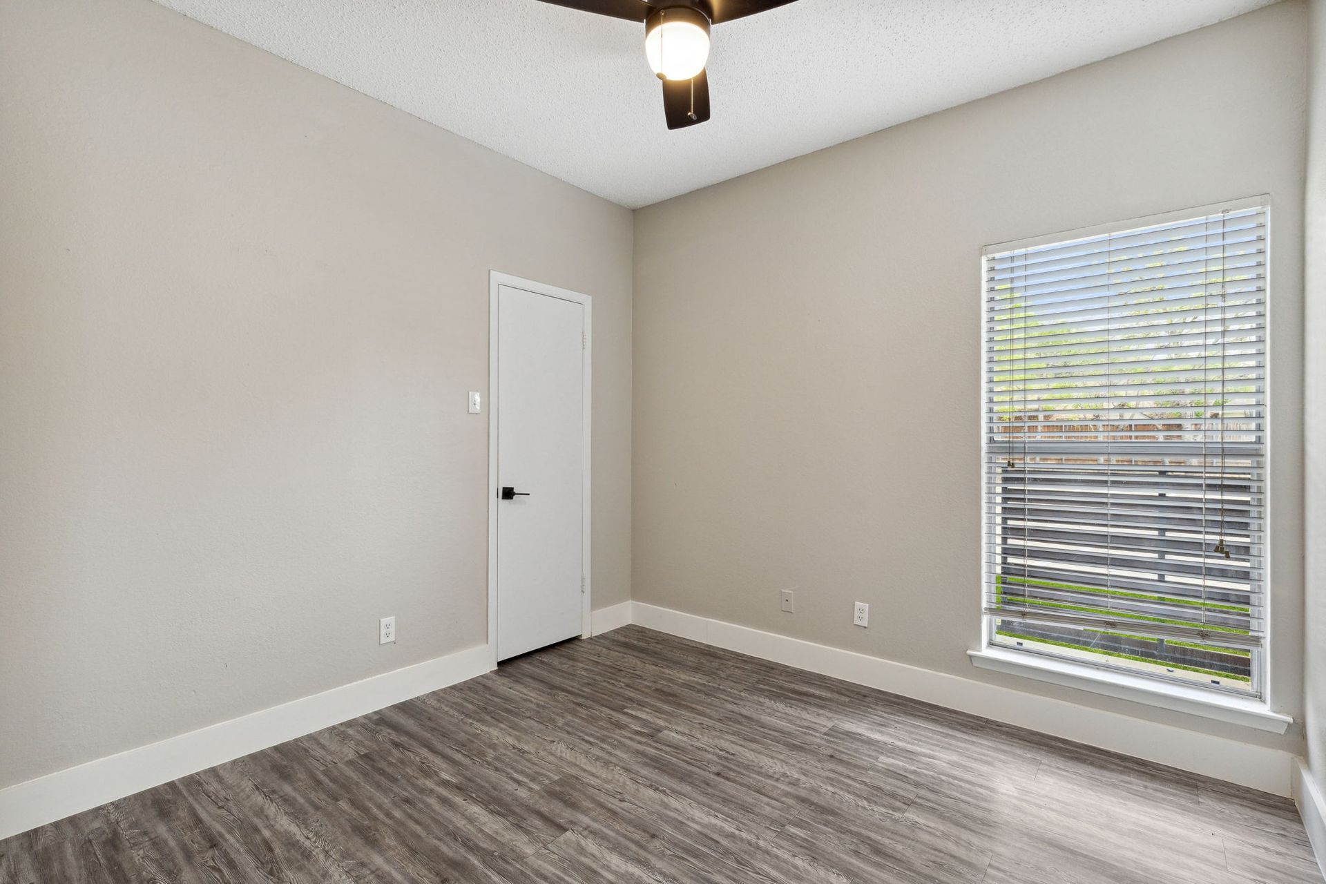 Empty bedroom with gray walls, wood-look floor, white trim, closed door, and window with blinds.