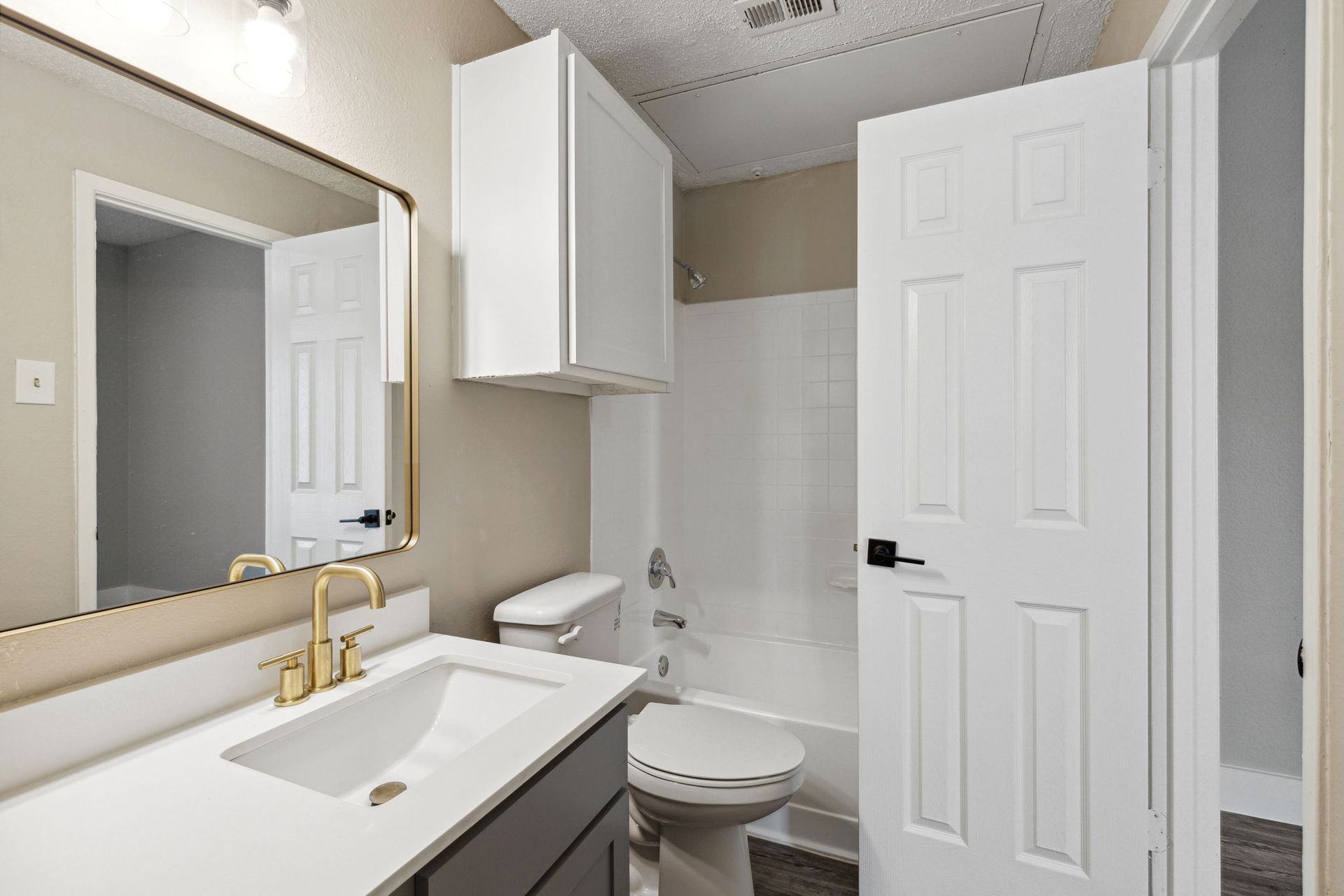 Bathroom with white vanity, toilet, and shower. Gold faucet and framed mirror. White door on the right.