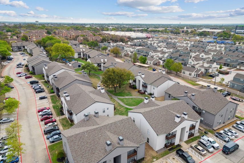 Aerial view of apartment complex with gray roofs and parked cars, against a green landscape and blue sky.