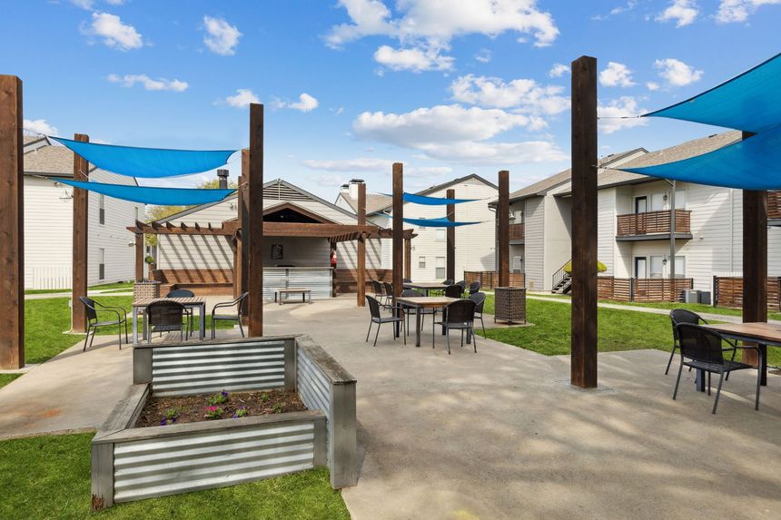 Outdoor seating area with blue shade sails, tables, chairs, and a raised garden bed.