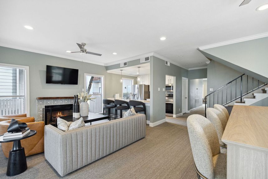 Fireplace, stairs, and kitchen access. Light gray walls, neutral furniture, and carpet.