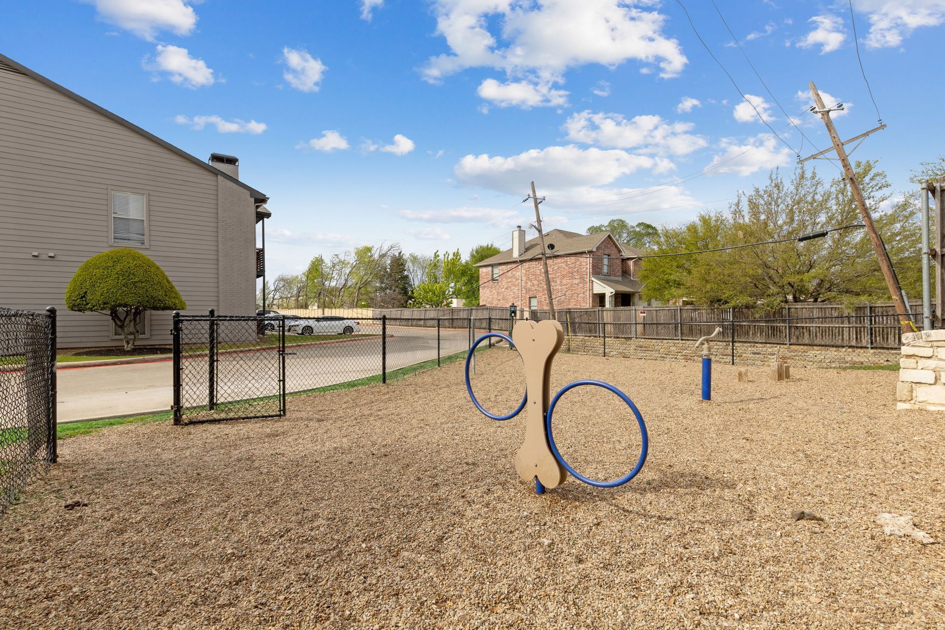 Dog park with wooden chips, agility equipment, fenced in, apartment building, cloudy sky.