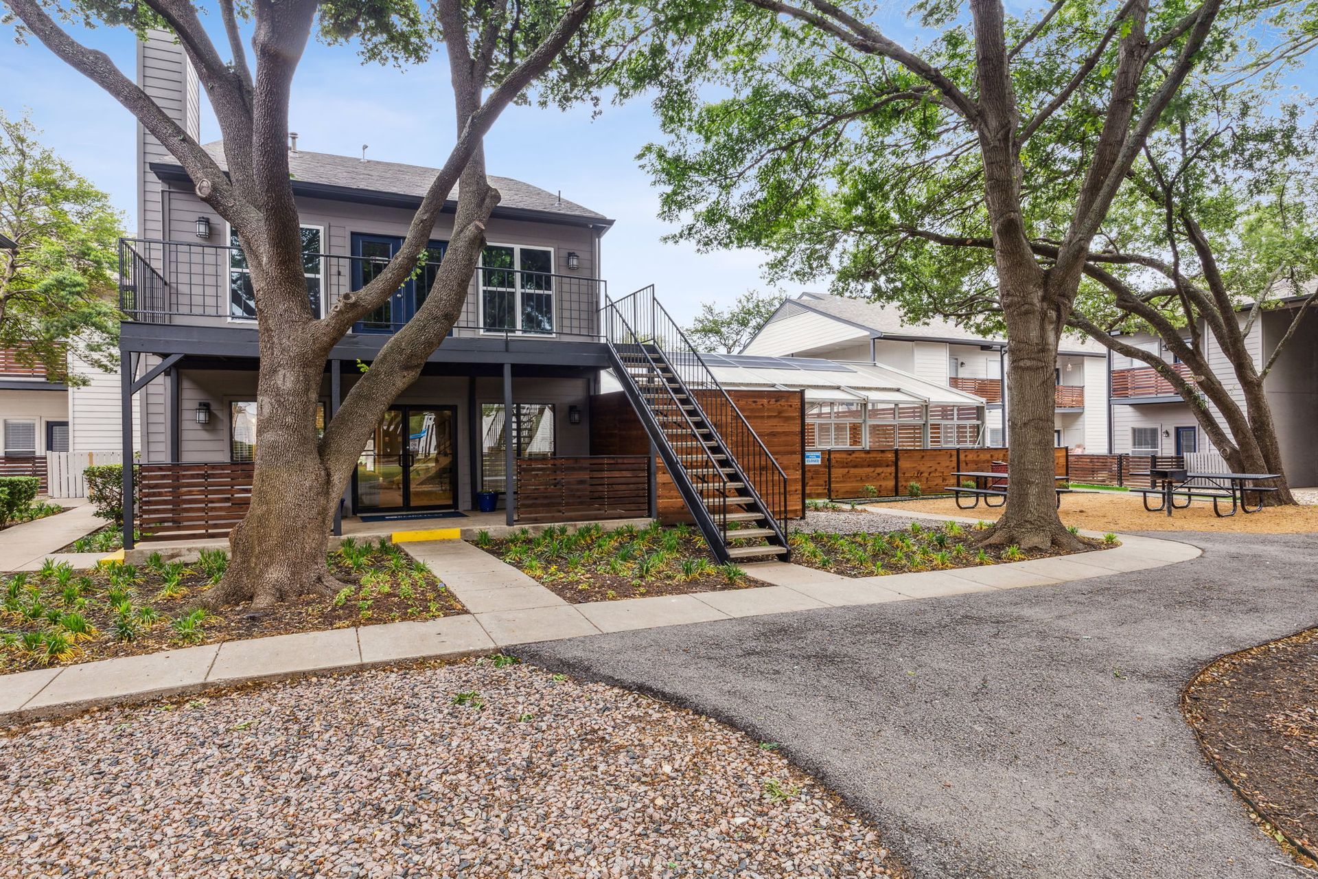 Two-story gray building with metal staircase and black railing. Trees frame the building with gravel path.