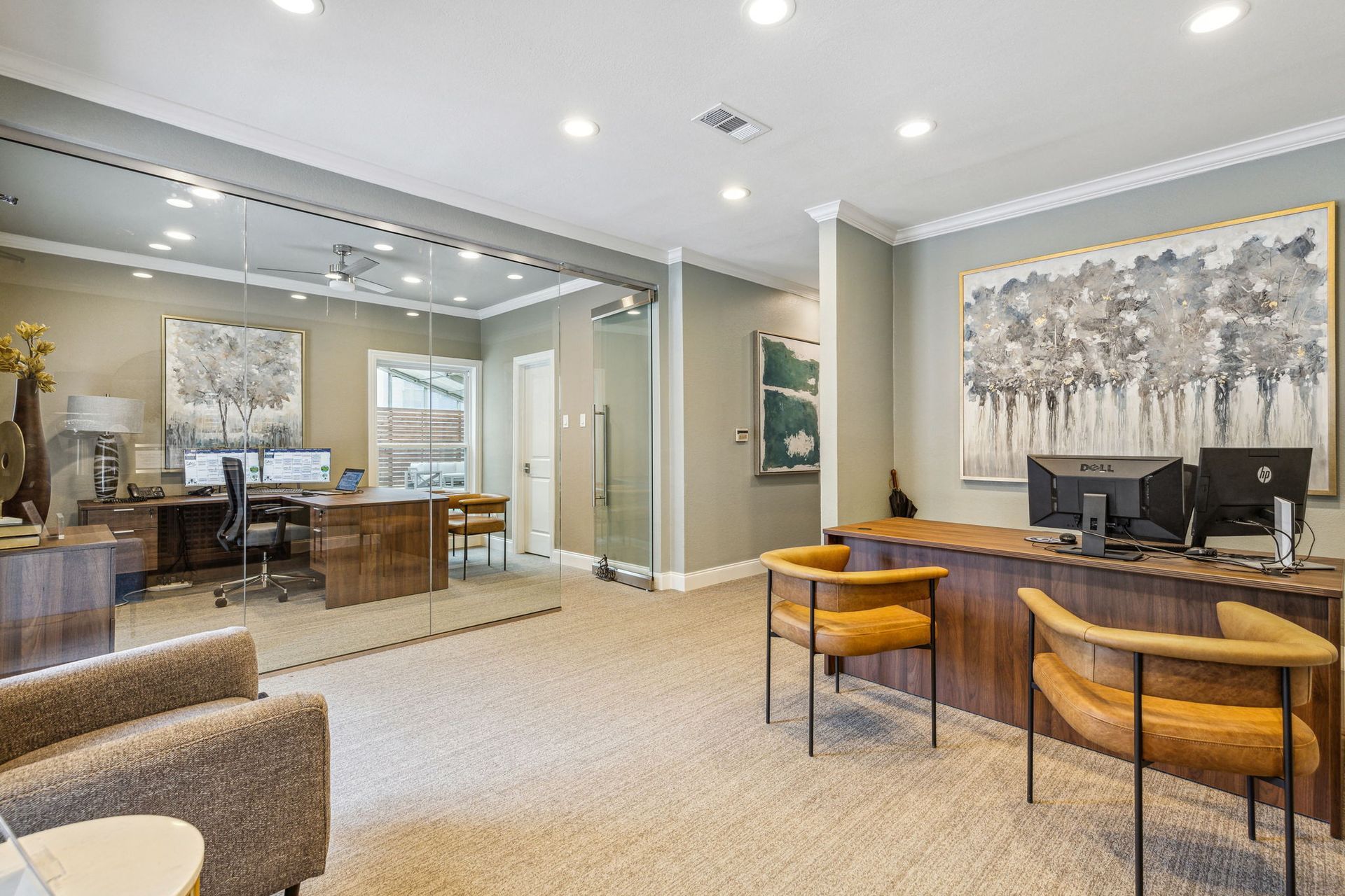 Office interior with desks, chairs, artwork, and glass-walled workspaces. Beige carpet, neutral walls, and modern decor.