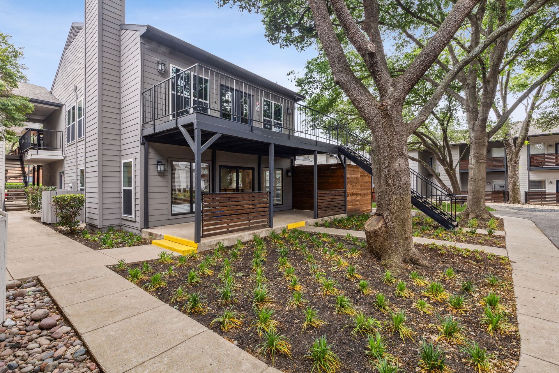 Apartment building with a wooden balcony and landscaping, trees line the walkway on a sunny day.
