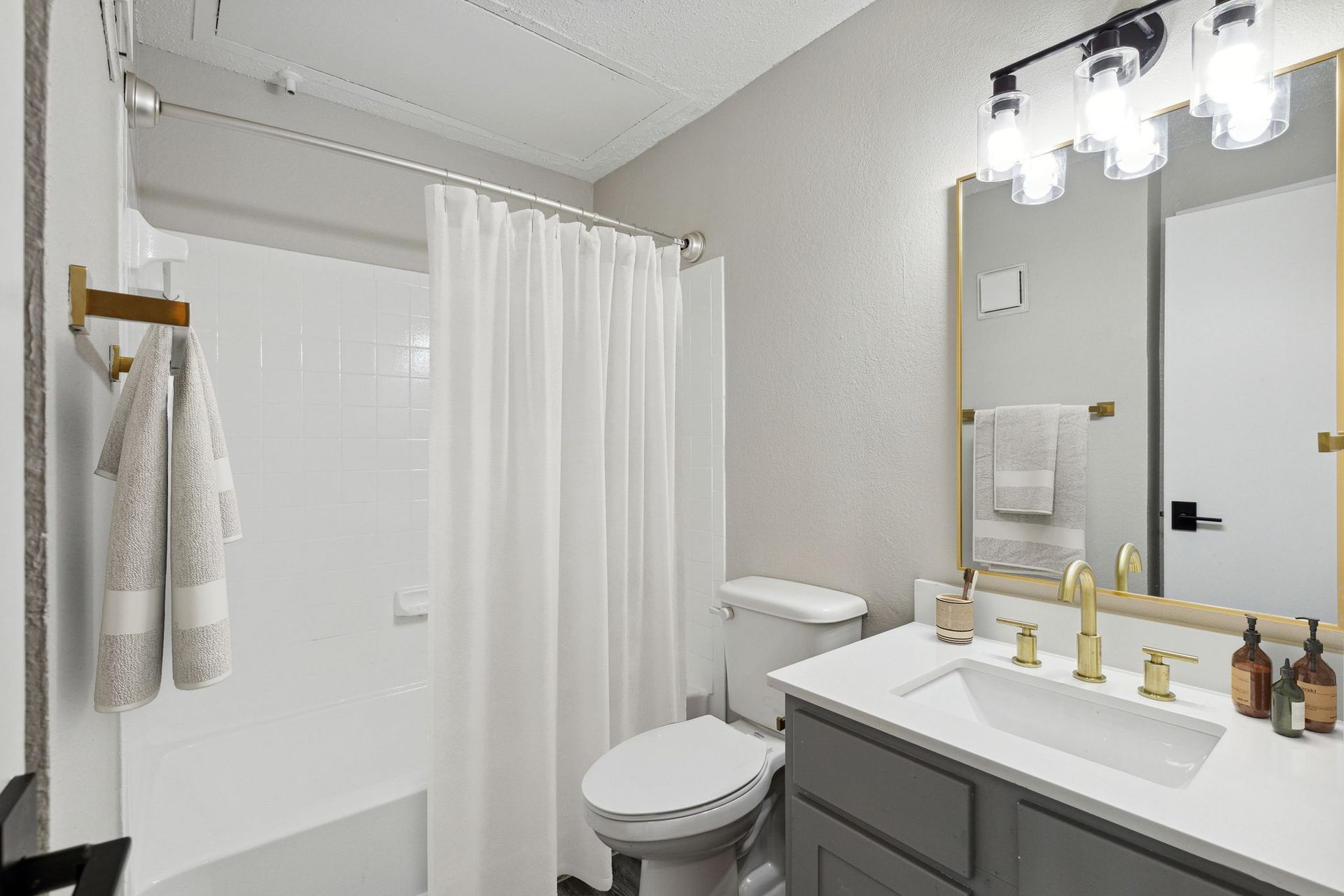 Bathroom with white tub, toilet, and sink. Gray vanity, gold fixtures, and bright overhead lights.