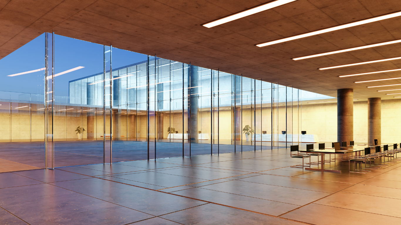 Modern glass-walled hall with wooden ceiling, tiled floor, and a few tables and chairs at sunset
