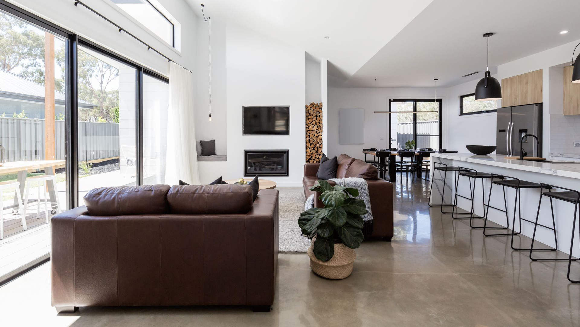 Bright open-plan living room with large windows, brown sofa, and kitchen island with barstools