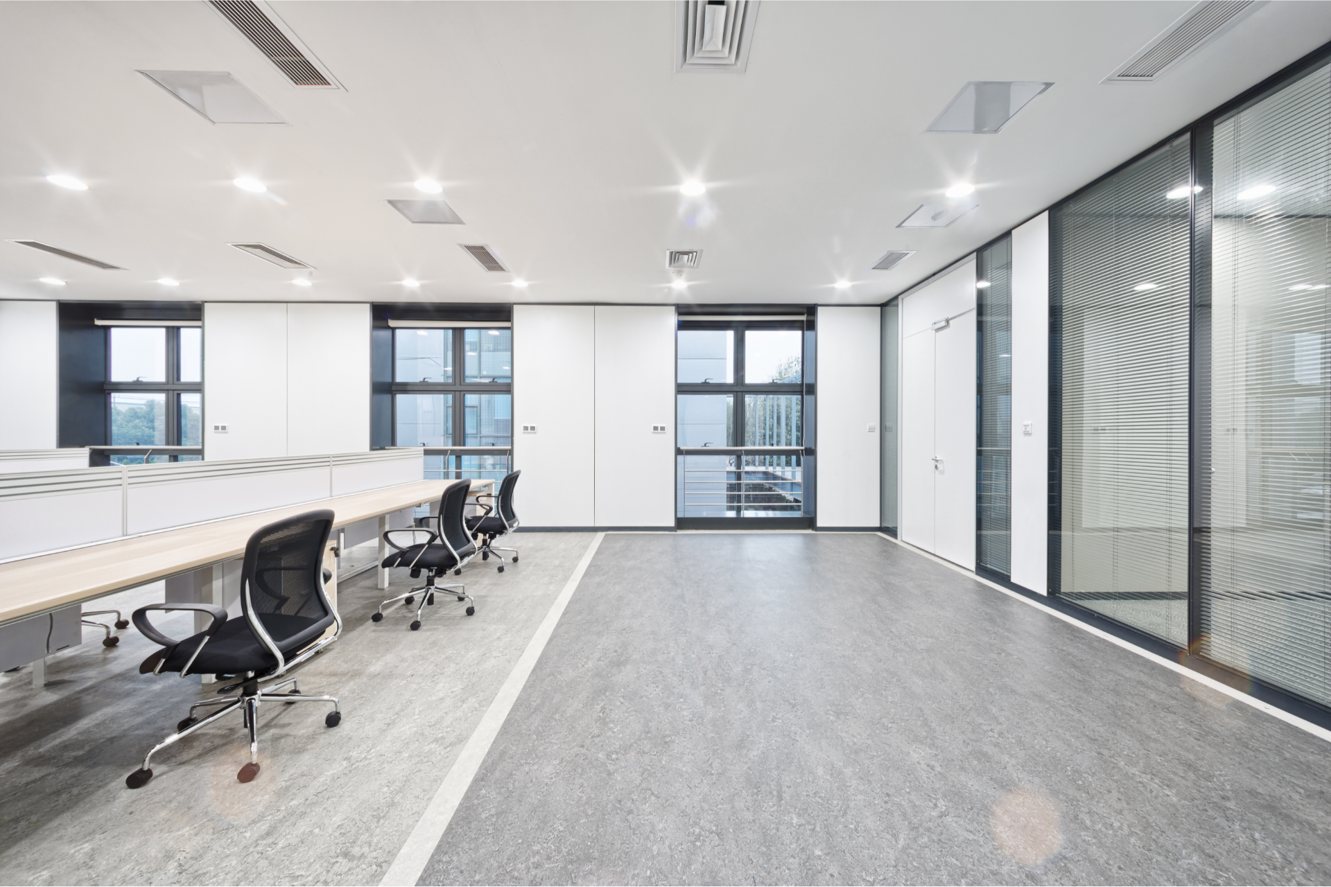 Modern office interior featuring a long wooden desk with black chairs, gray flooring, and glass-partitioned rooms.