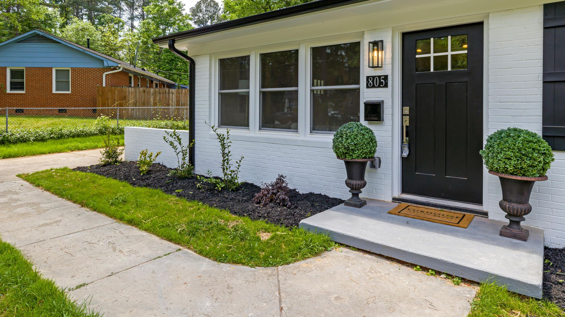 White house entry with black door, potted topiaries, and a concrete walkway in a suburban neighborhood