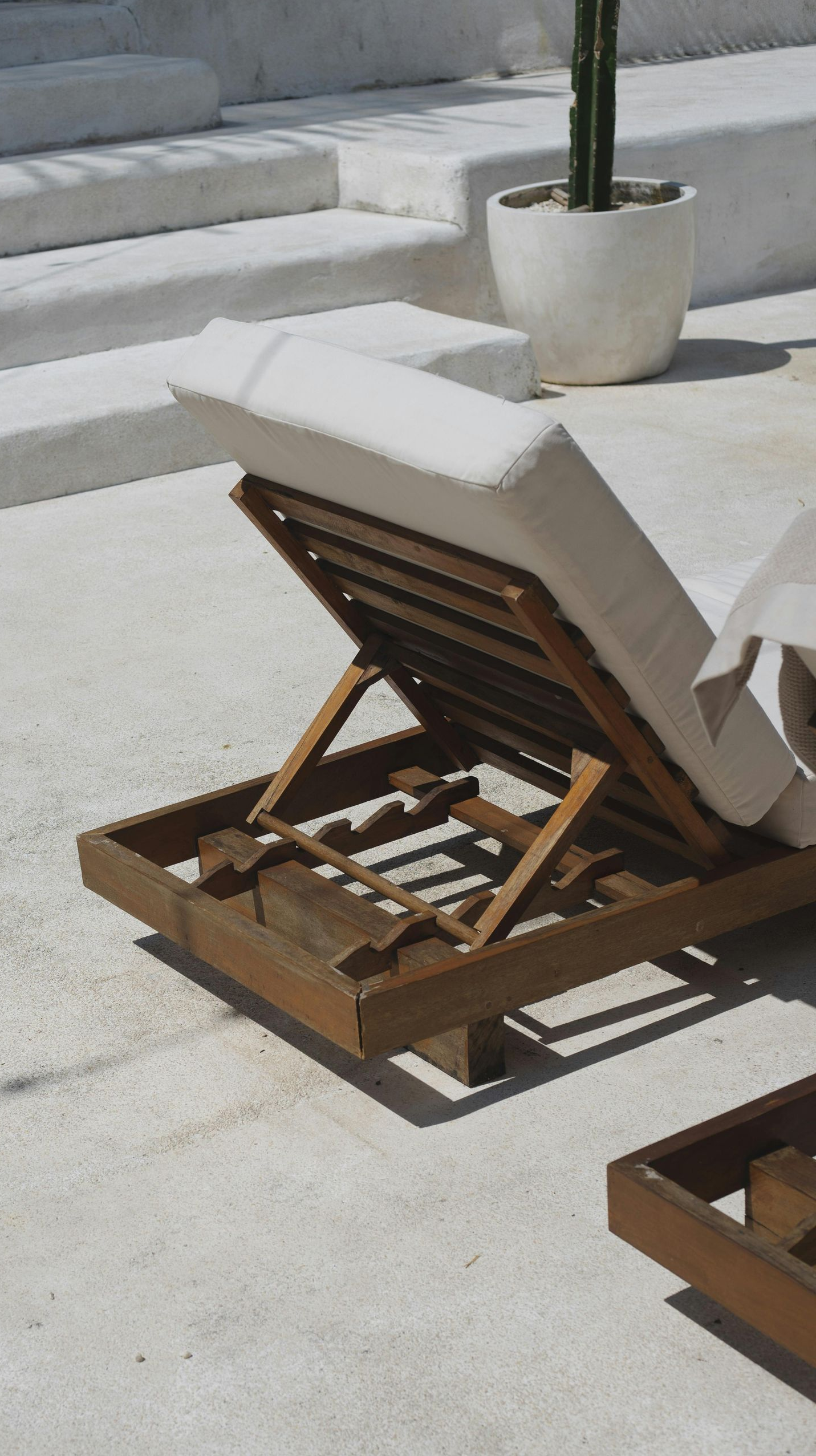 A wooden outdoor lounge chair with white cushion is open on a sunlit patio near steps and a potted plant.