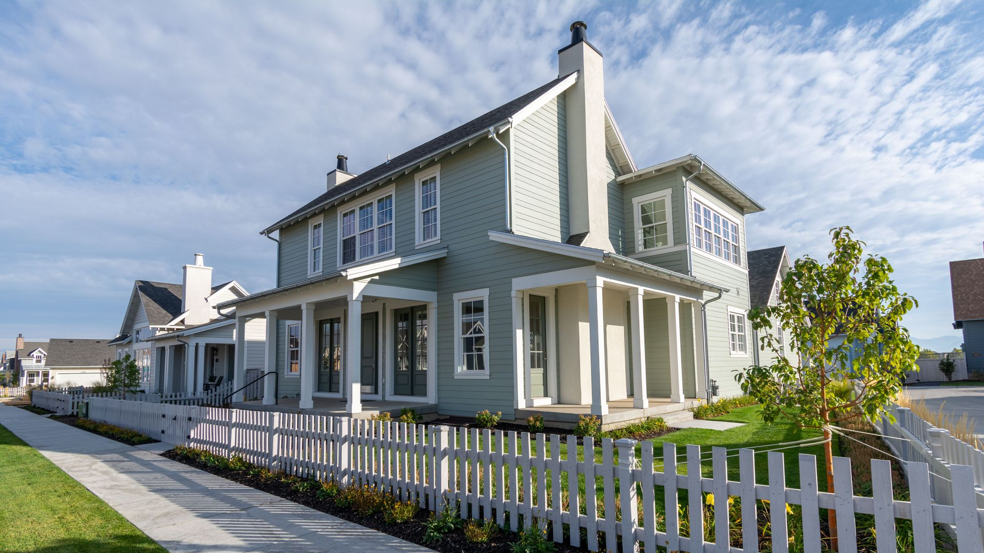 White two-story house with porch and white picket fence along a sidewalk on a sunny street