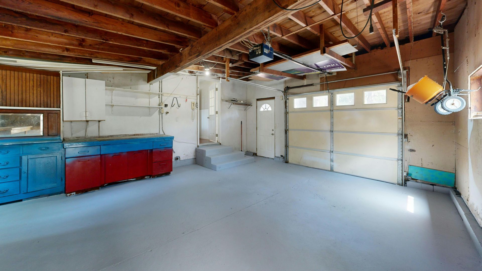 Empty garage interior with workbench, white cabinets, exposed beams, and concrete floor