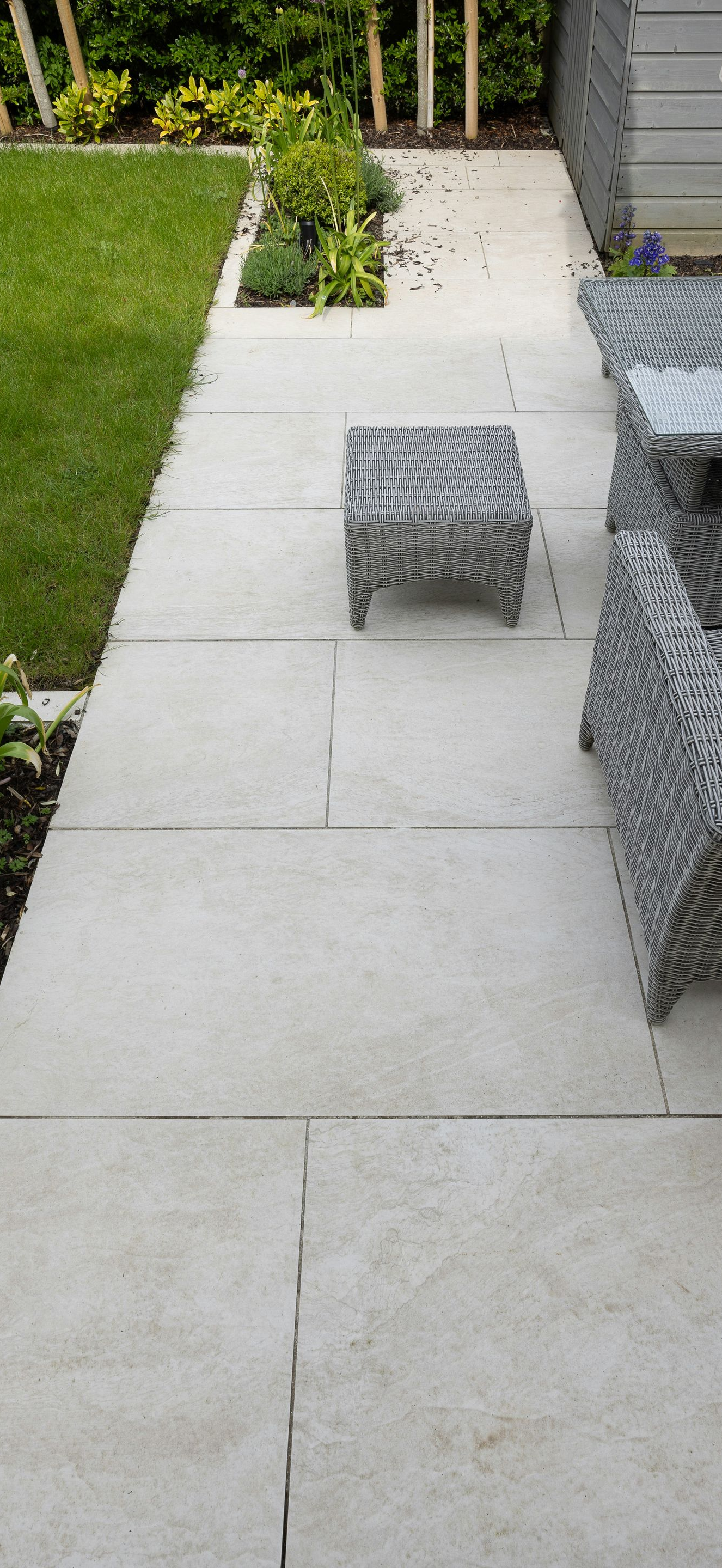 Patio walkway with white pavers, a small woven stool, and outdoor seating beside a lawn and garden.