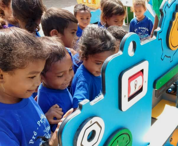 Children in blue shirts examine a blue play structure with gears and shapes outdoors.