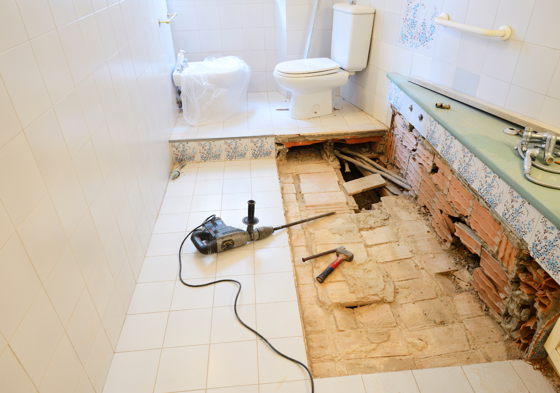 Bathroom under renovation; floor tiles removed, exposing brick and plumbing. A jackhammer and tools lie on the floor.