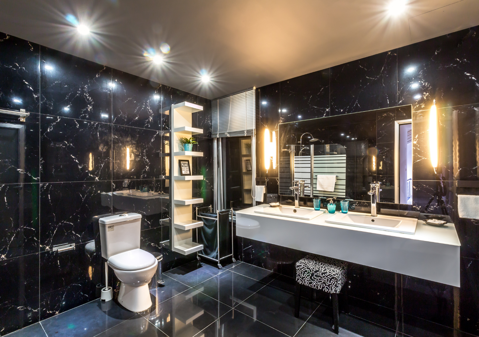 Luxurious bathroom with black marble walls, white fixtures, and modern lighting. Includes a toilet, sink, shelving unit, and a stool.