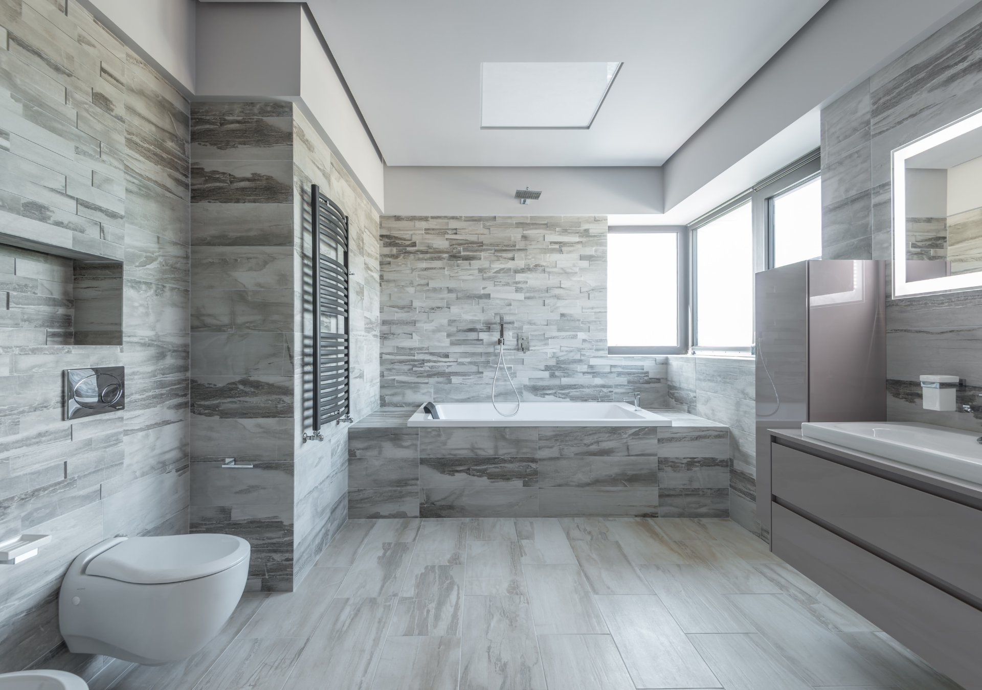 Modern bathroom with gray stone tile, a white toilet, a built-in bathtub, and a large window.