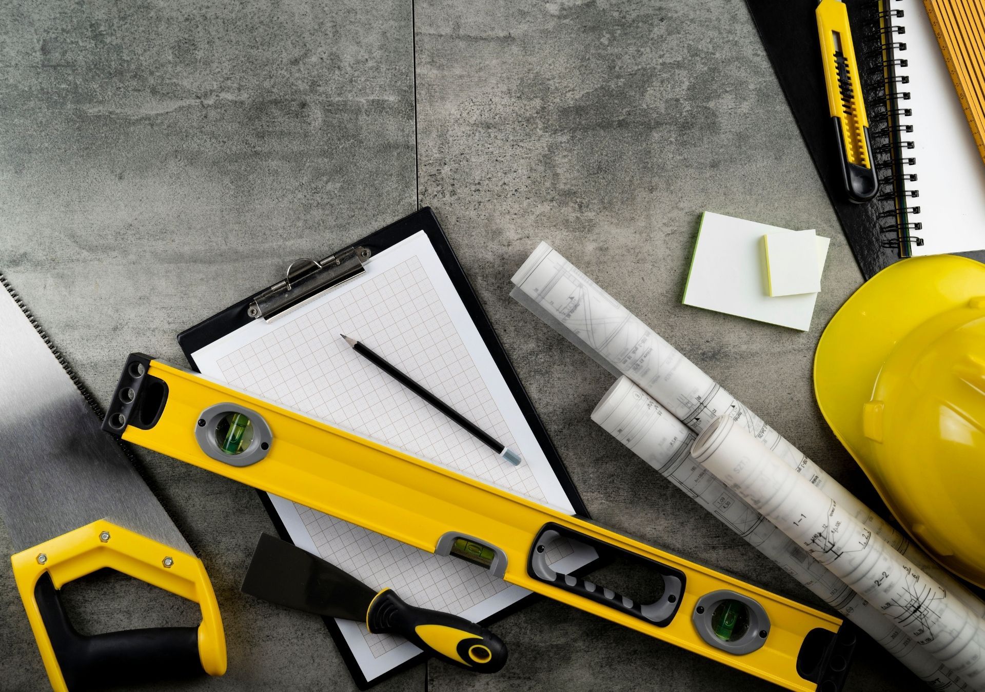 Construction tools: level, blueprints, clipboard, saw, hard hat, on a grey surface.