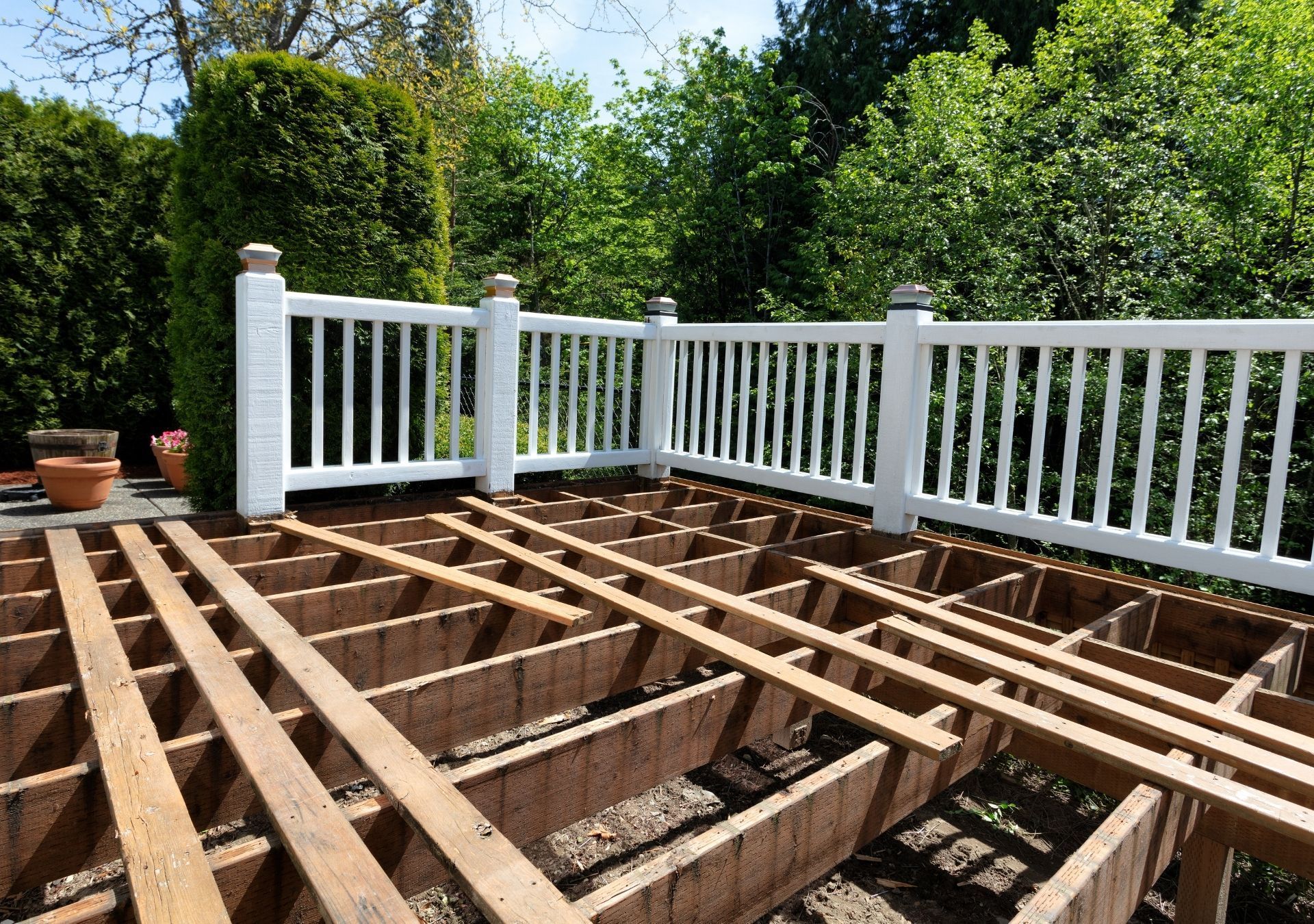 A partially disassembled deck with a white railing in front of a green tree backdrop.