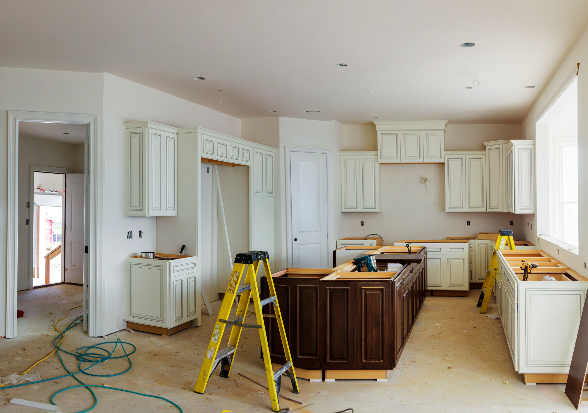 Kitchen renovation in progress with light cabinets, a dark island, and a yellow ladder. Wires and tools on the floor.