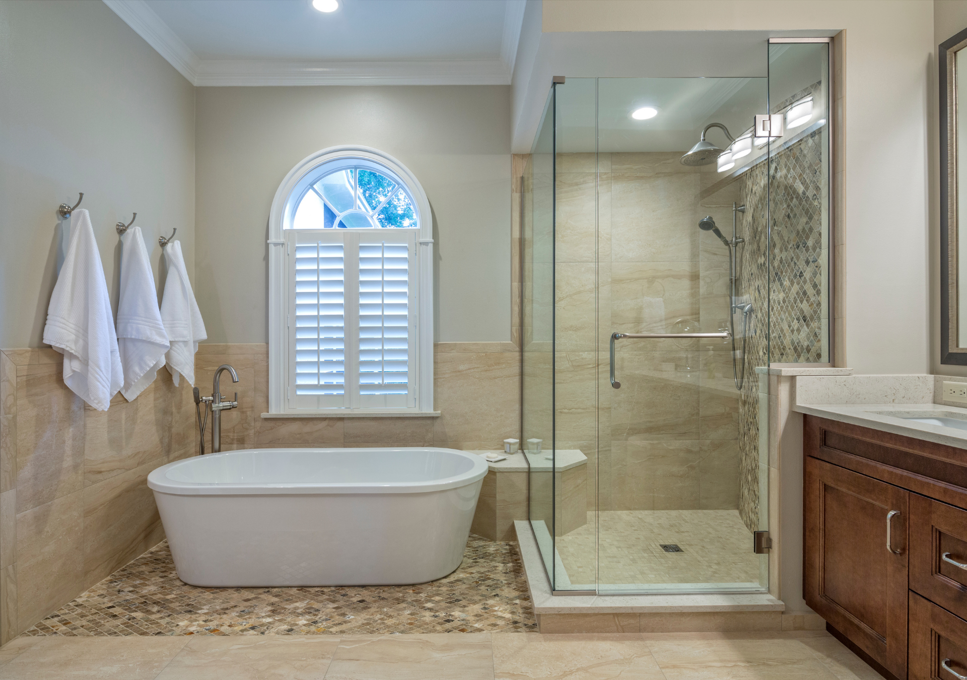 Luxurious bathroom with a freestanding tub, glass shower, and shuttered window. Beige walls, stone floor, and dark wood vanity.