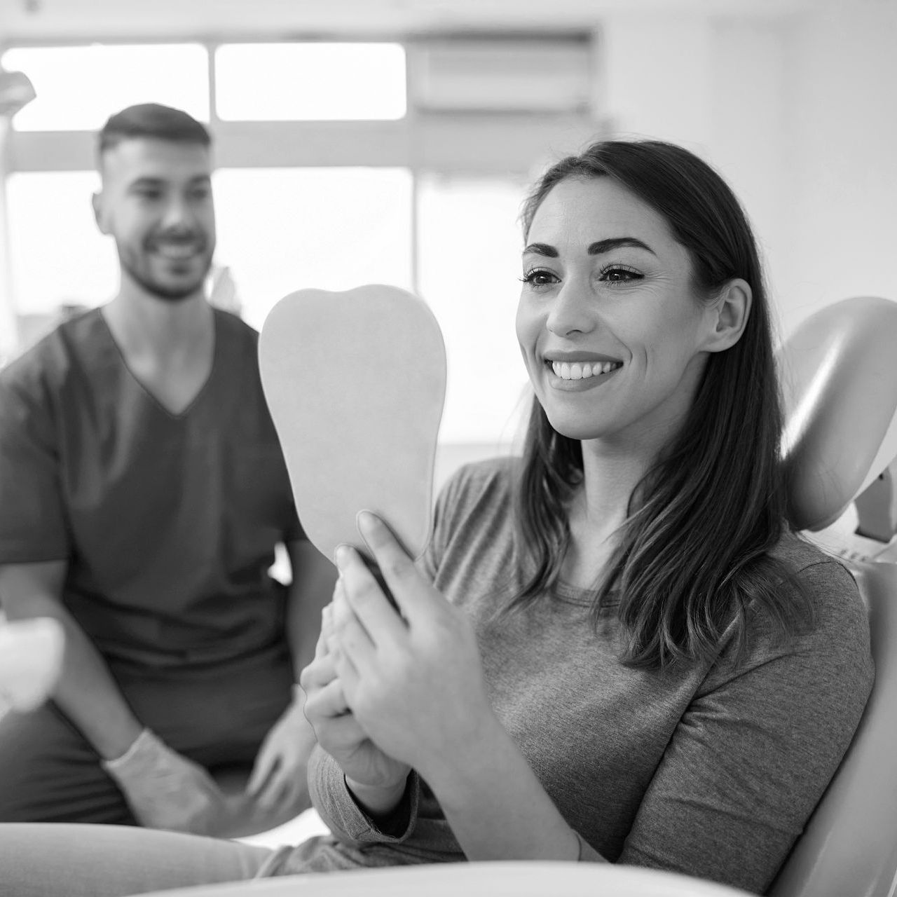 A woman is sitting in a dental chair looking at her teeth in a mirror.