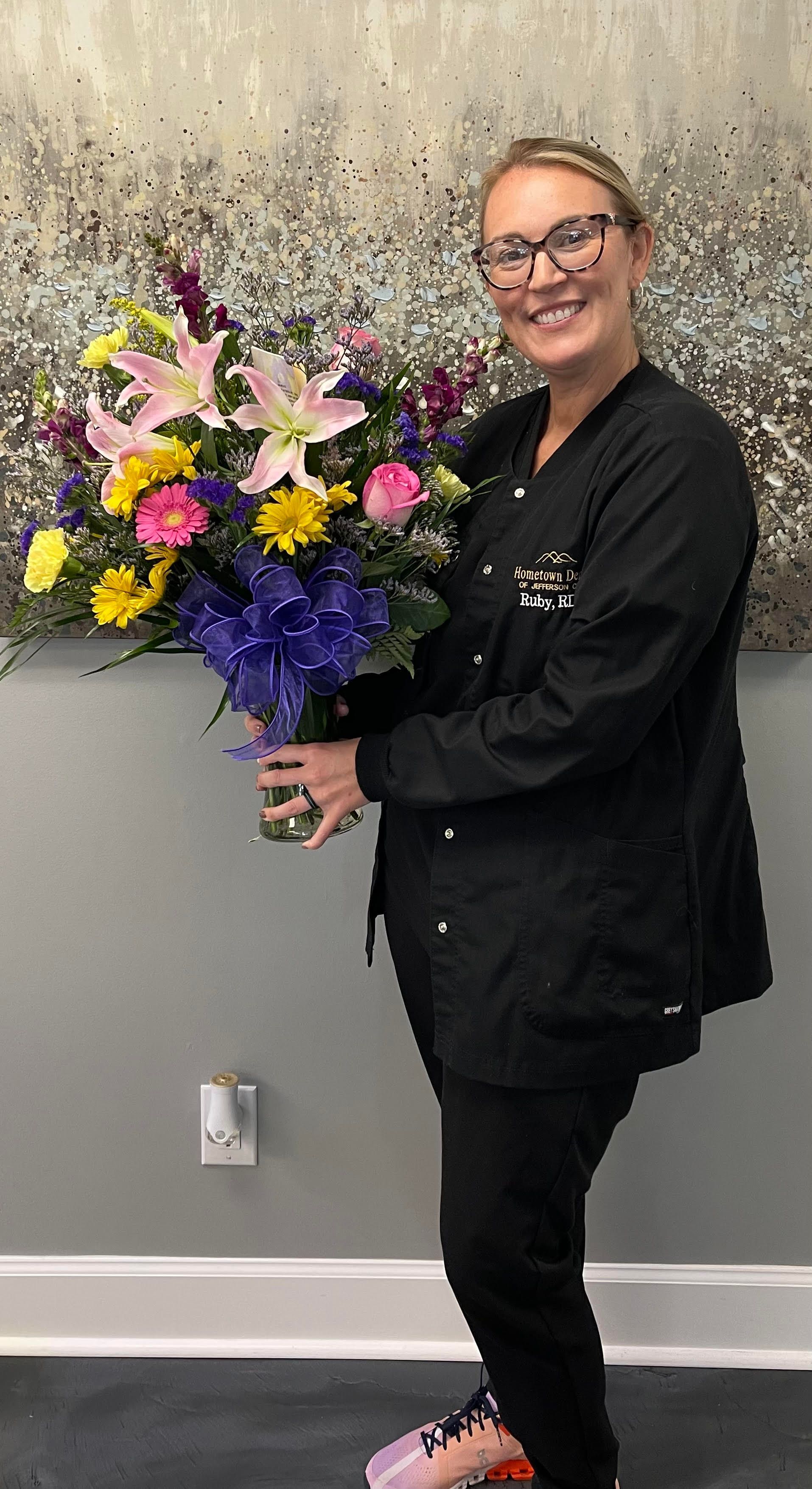 Woman in black scrubs holding a large bouquet of colorful flowers, smiling. Standing in front of a gray wall.