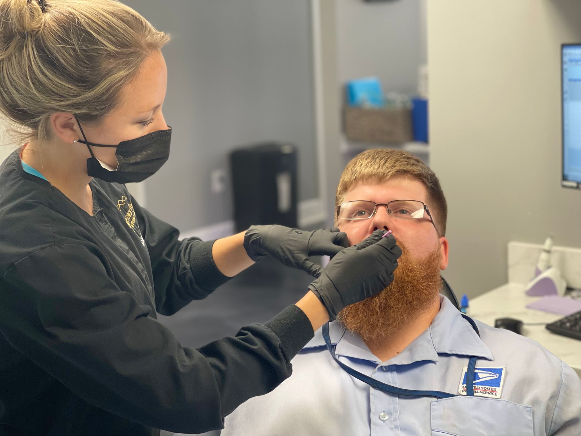 Dental assistant wearing a mask and gloves, examining a man's mouth. Office setting, seated.
