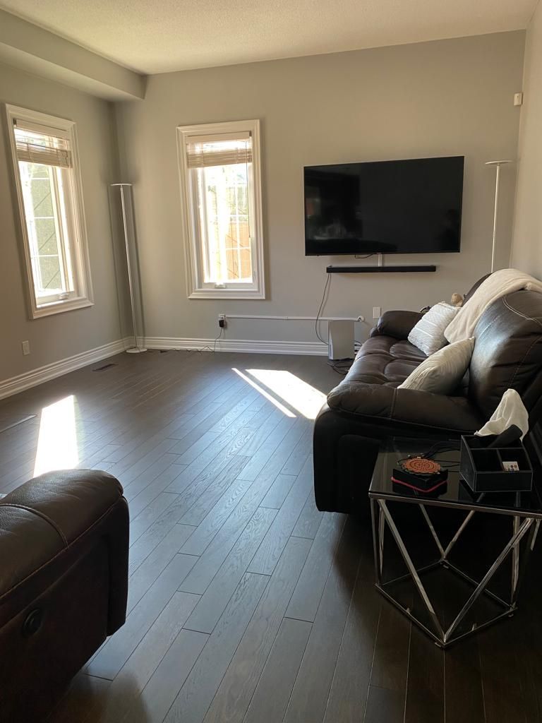 Living room with dark wood floors, large TV, brown leather sofa, and two windows.