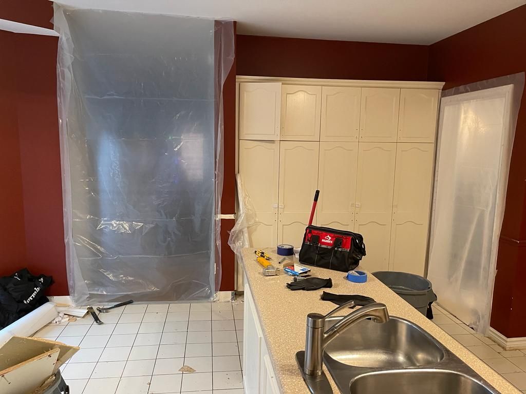 Kitchen under renovation: plastic sheeting, red walls, white cabinets, tools on counter, tiled floor.