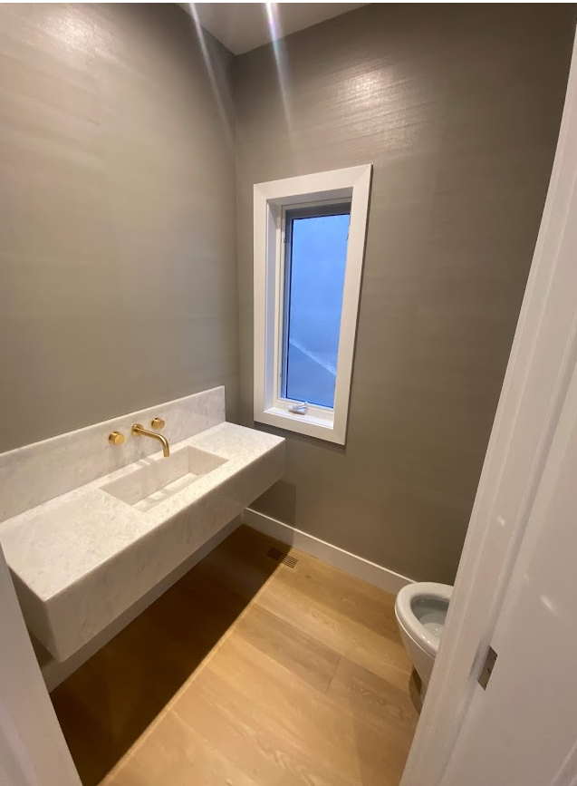 Modern bathroom with floating vanity, gold faucet, window, toilet, and light-colored wood floor.