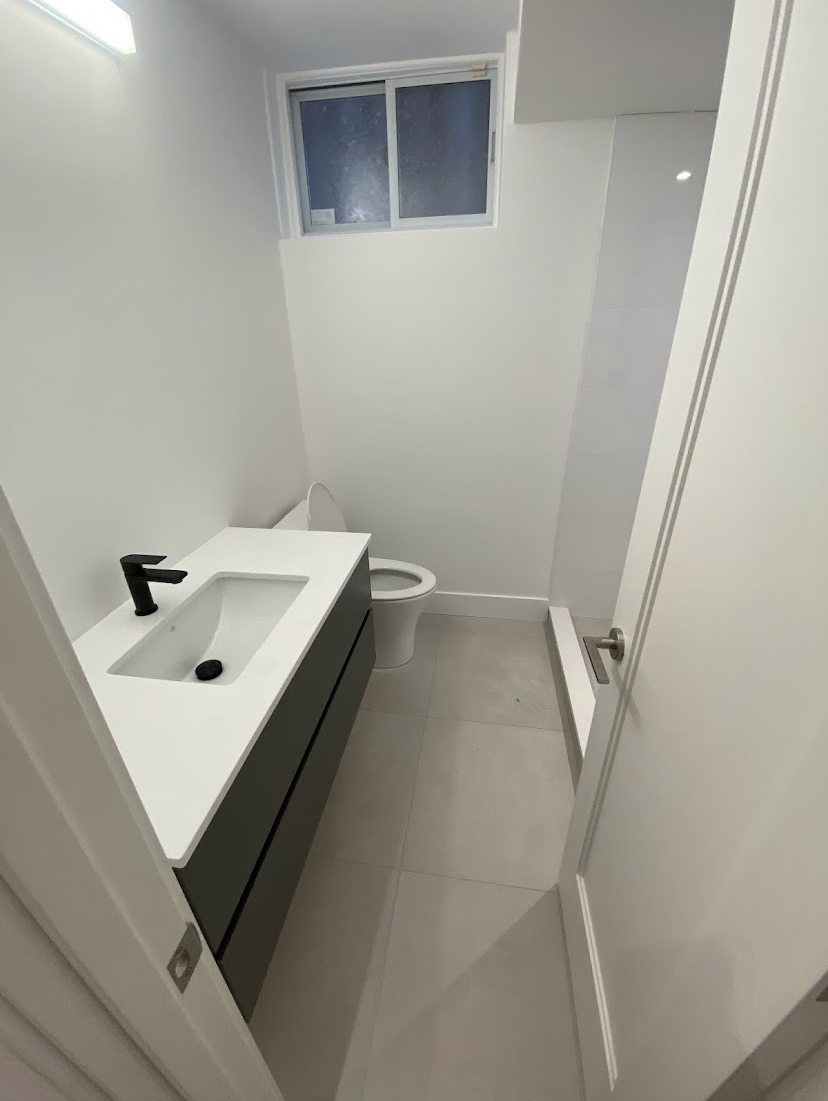 Small, modern bathroom with a gray vanity, white toilet, and window; white walls and floor.