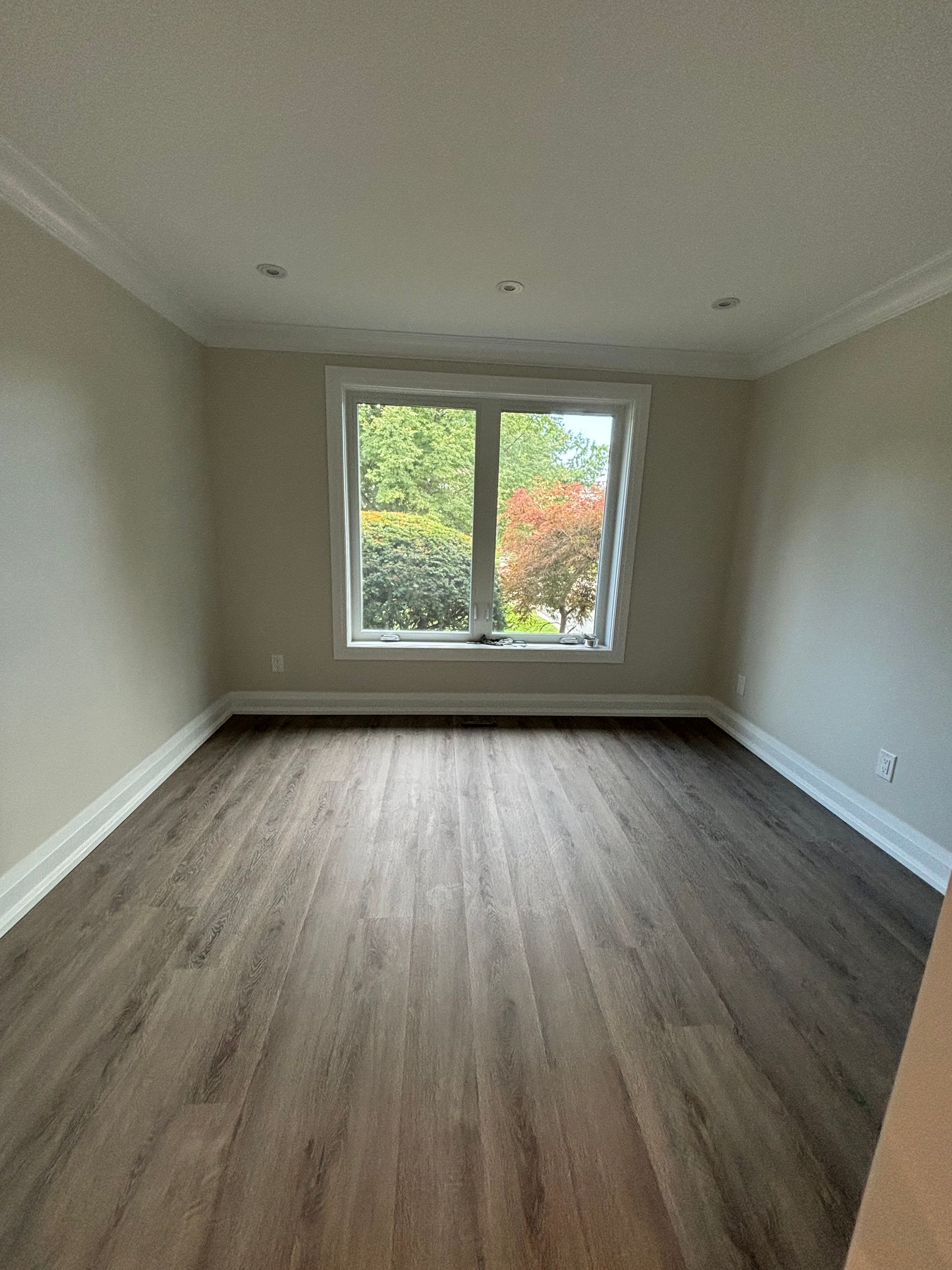 Empty room with dark wood-look flooring, beige walls, white trim, and a window with a view of greenery.