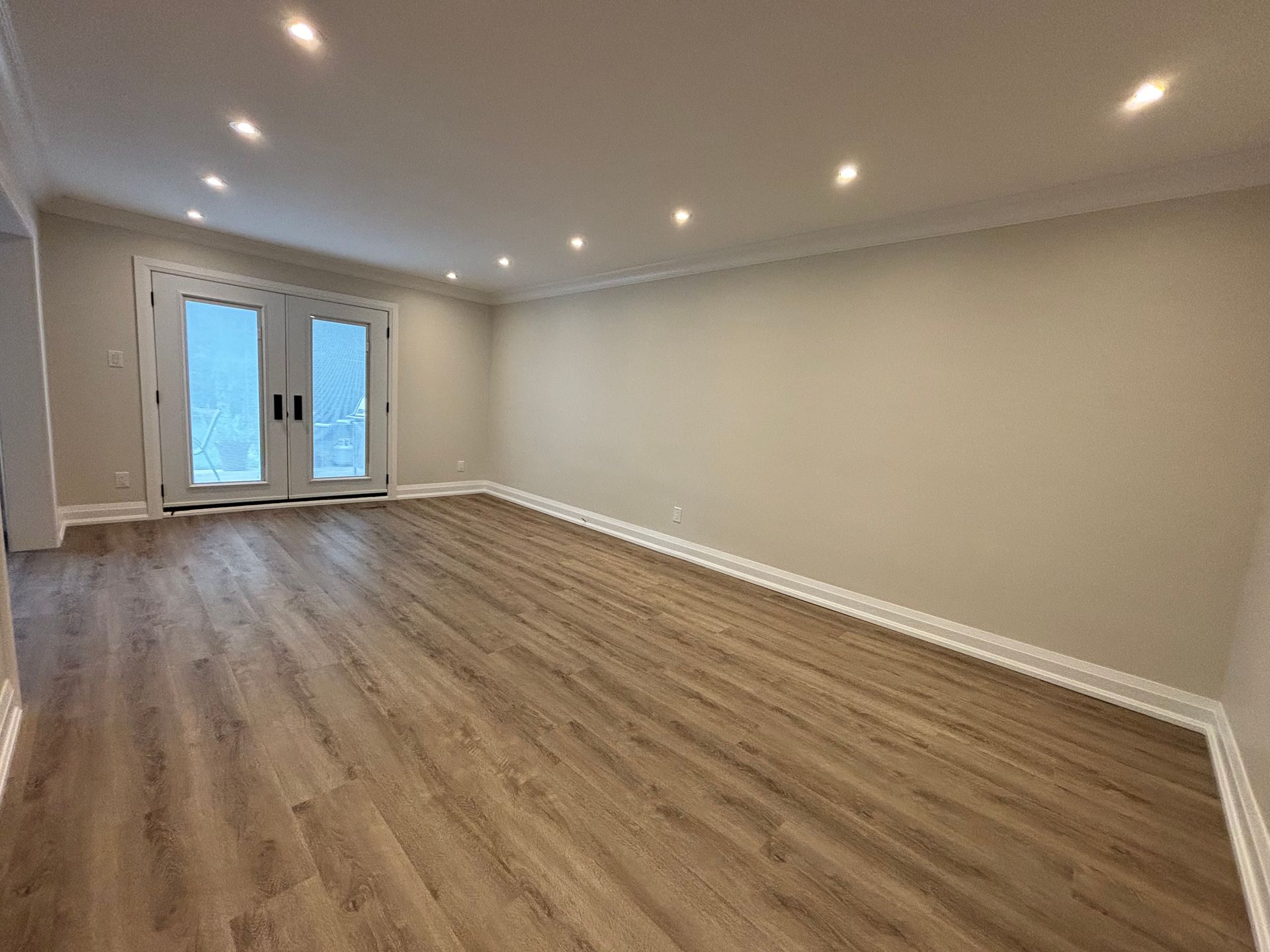 Empty room with light wood flooring, beige walls, recessed ceiling lights, and double doors.