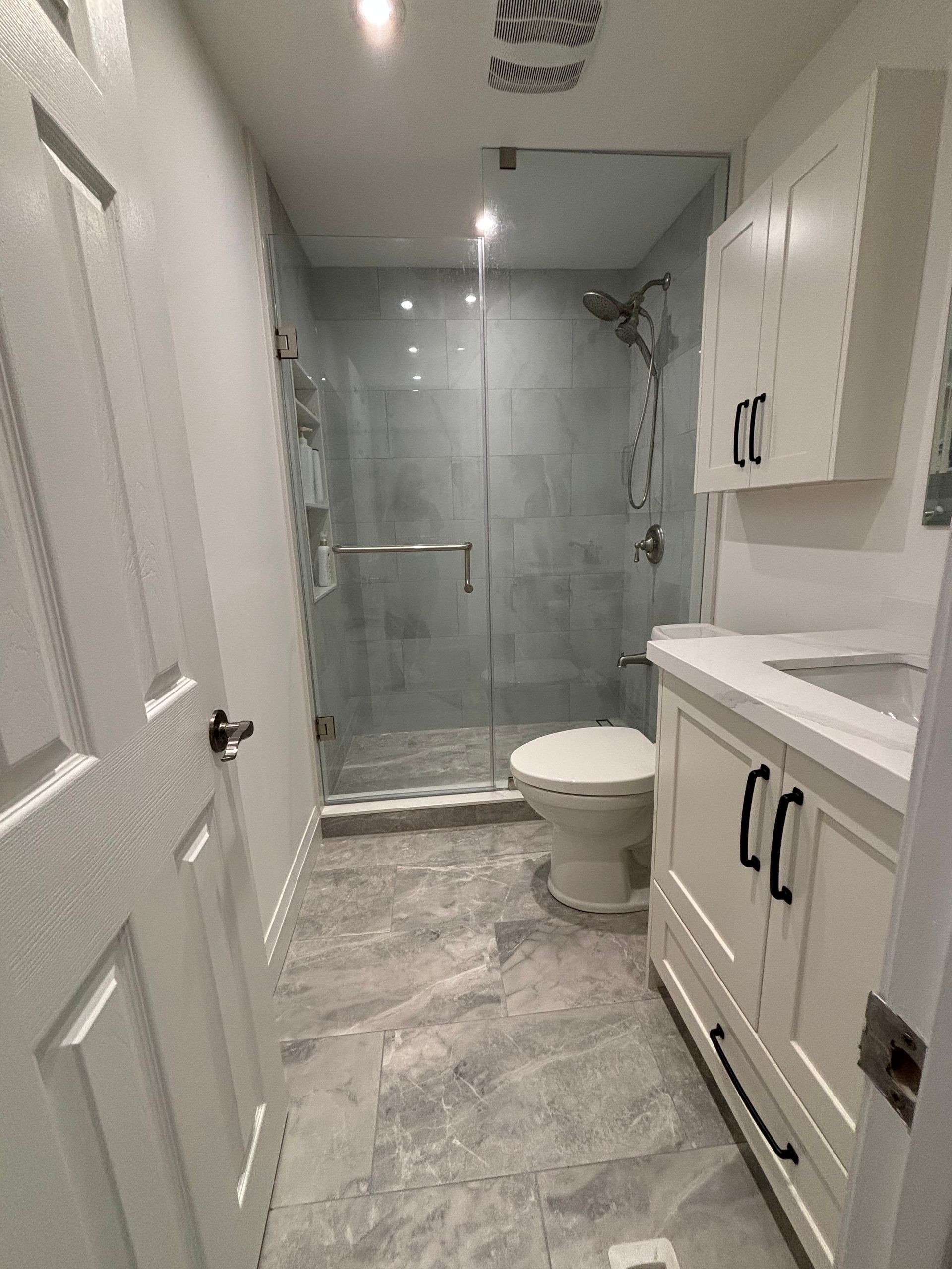 Small modern bathroom with gray tile floor, glass shower, white vanity, and toilet.