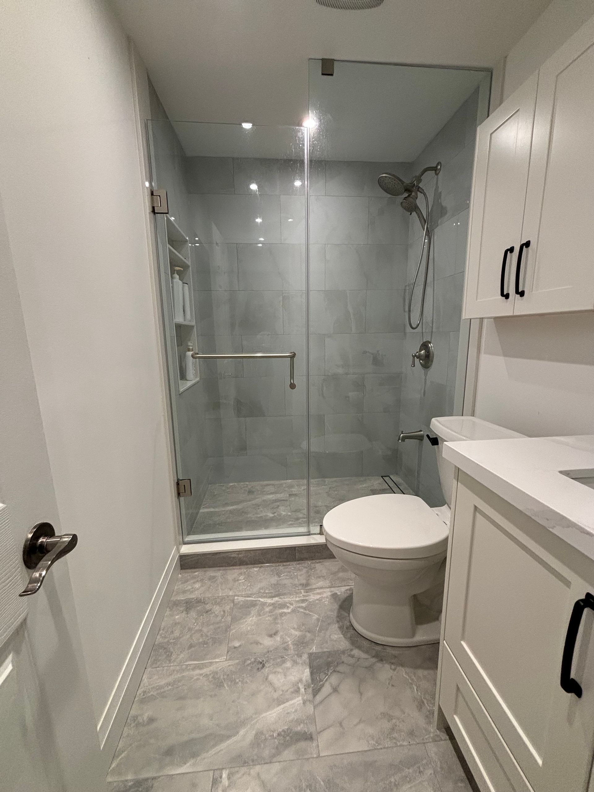 Modern bathroom with gray tiled floor, glass shower, white cabinets, and toilet.