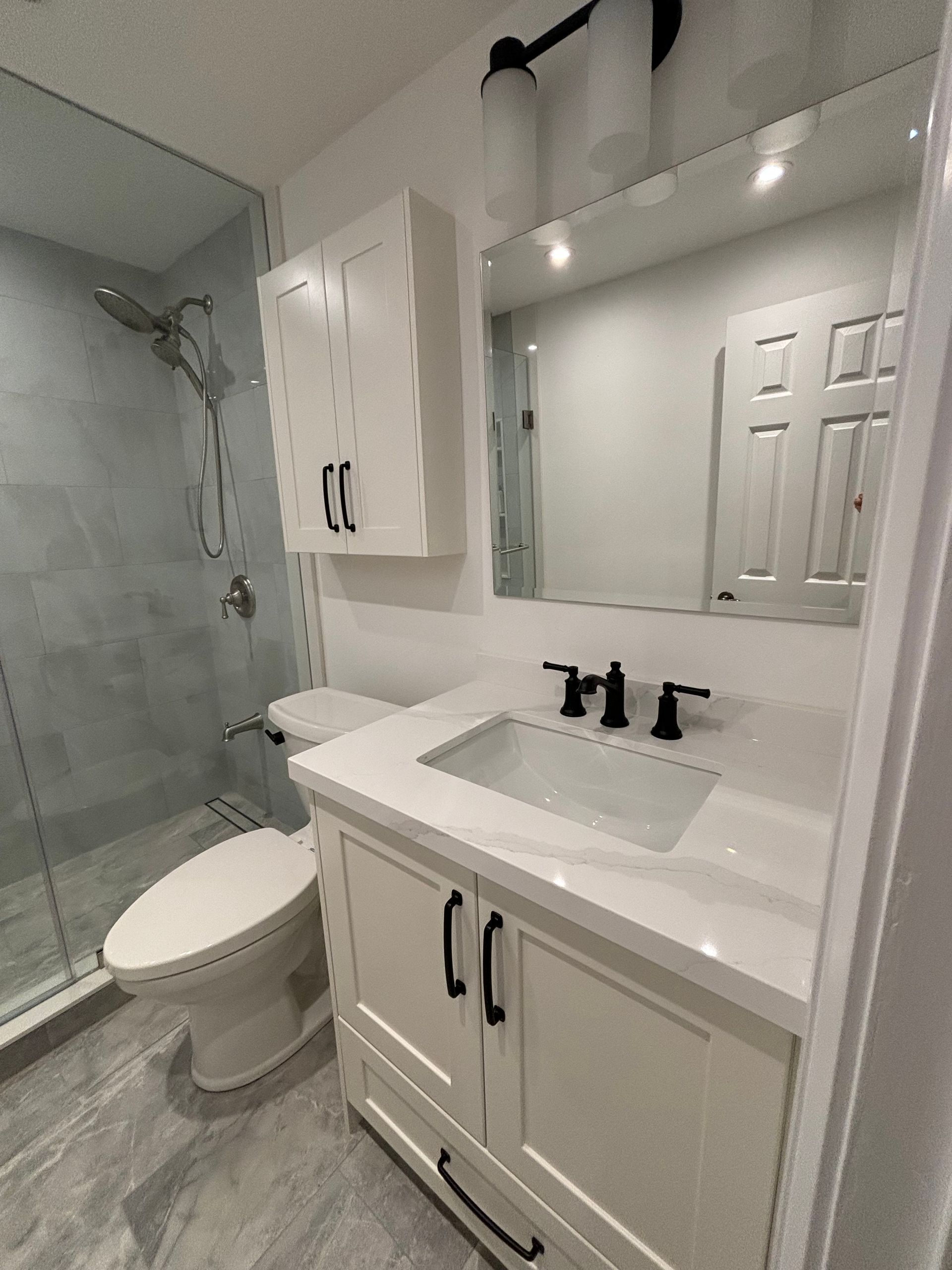 White bathroom with shower, vanity, toilet, and mirror. Black hardware and white cabinets. Gray tiled floor.
