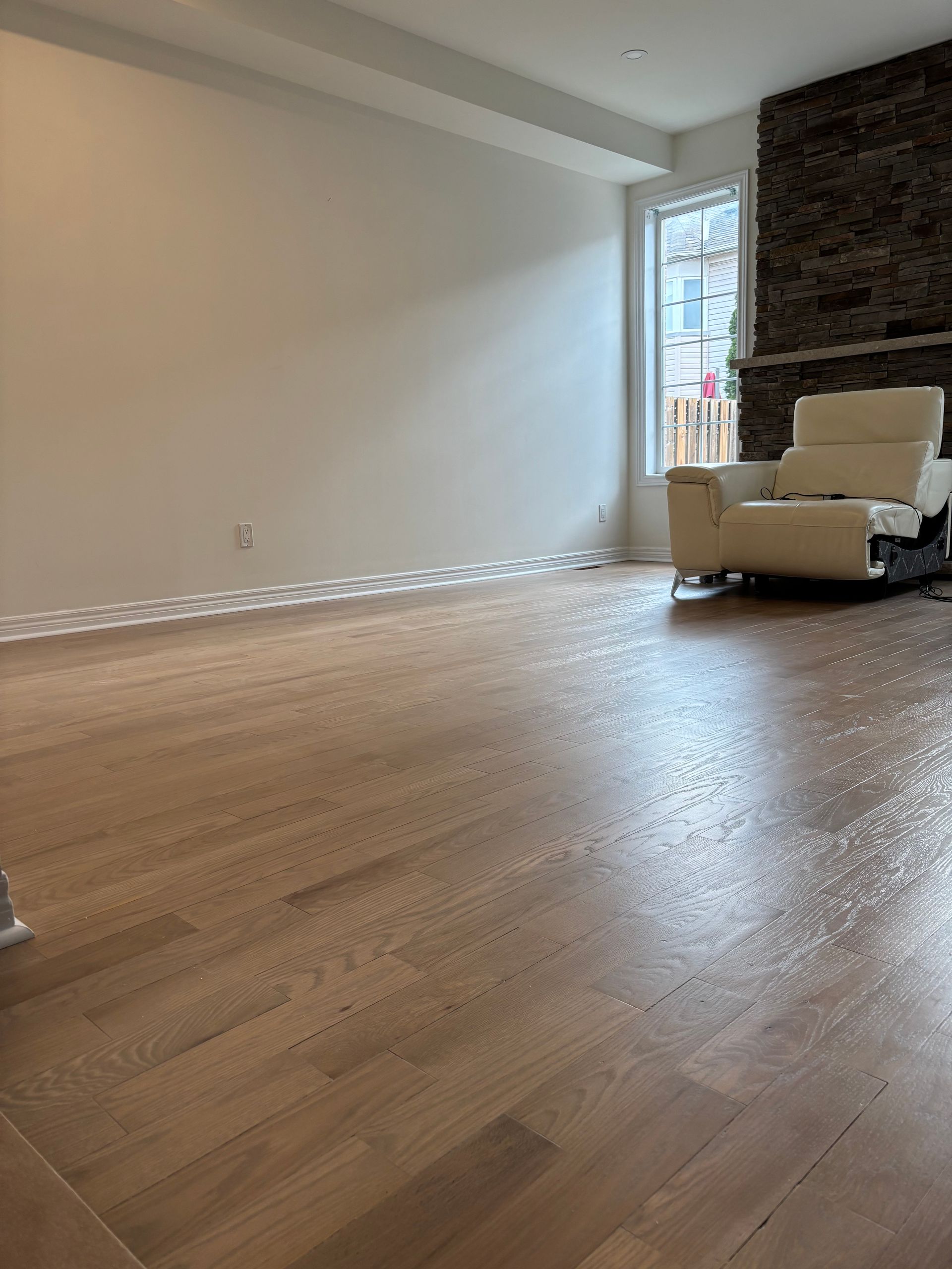 Hardwood floor in a room with a white wall, window, and a white chair near a brick fireplace.