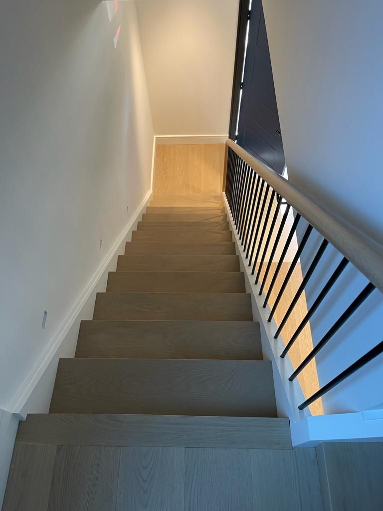 Staircase with light-colored wooden steps and railing with black vertical bars. Sunlight streams in from the top.