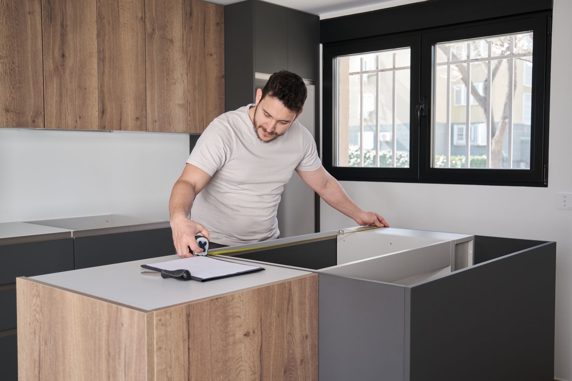 Man measuring cabinets in a modern kitchen, with clipboard and window.